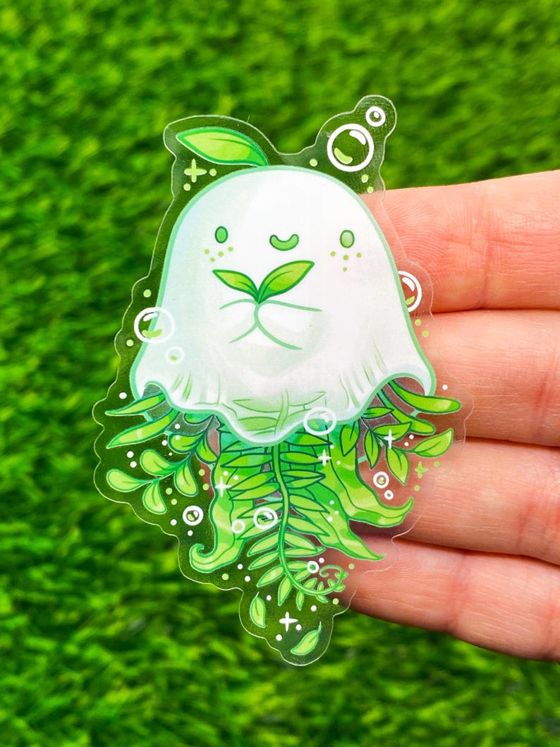 May include: A clear sticker featuring a whimsical white ghost-like character with green fern-like details. The character has a smiling face with green leaves and is surrounded by bubbles and leaves. The sticker is held against a green background.