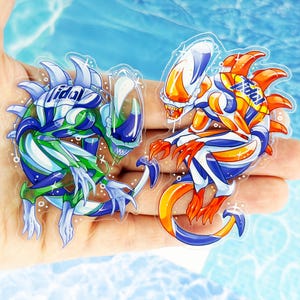 May include: Two clear acrylic stickers featuring stylized alien creatures with sharp teeth and tails. One creature is colored in shades of green, blue, and white, while the other is orange, blue, and white. Both stickers display the word Tidal.