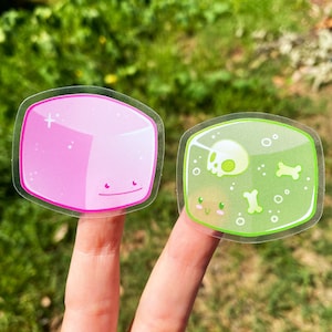 May include: Two clear stickers with cartoon characters. The first sticker is pink and features a square with a smiling face. The second sticker is green and features a square with a skull and crossbones inside.