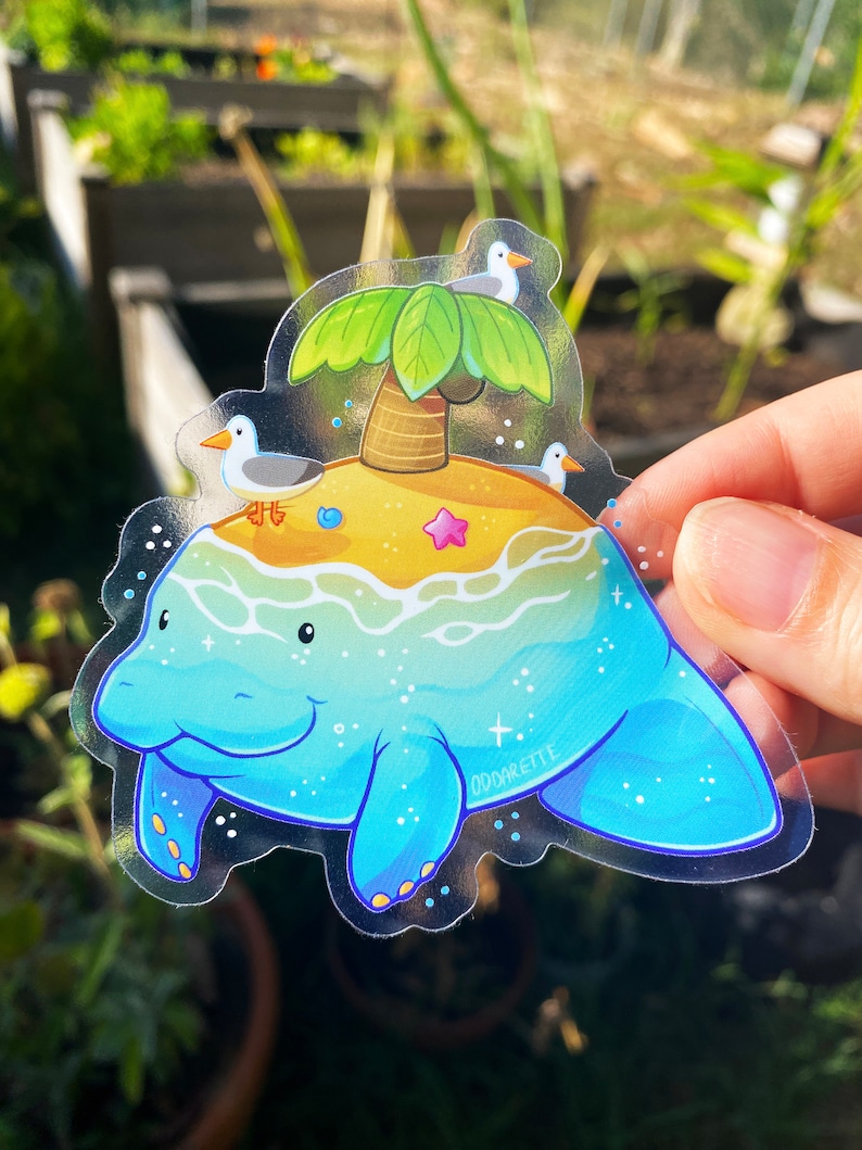 May include: A clear sticker featuring a cartoon manatee with a small island on its back. The island has a palm tree, a starfish, and three seagulls. The sticker has a blue, green, and yellow color scheme and the text "oddakettle" in the bottom right corner.
