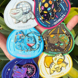 May include: A collection of colorful enamel pins featuring a cartoon character in various designs. Each pin is on a unique, decorative backing with the text "ODDARETTE". The pins showcase different color schemes and themes, including a disco ball and a lemon.