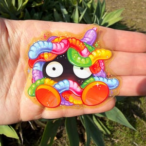 May include: A colorful sticker featuring a cartoon creature with a black face, large white eyes, and a variety of colorful worm-like appendages in shades of red, orange, yellow, green, blue, and purple. The sticker has a clear, glossy finish.