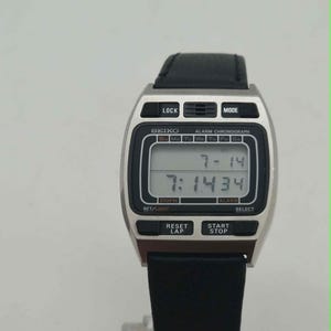 May include: A silver-tone digital Seiko alarm chronograph watch with a black leather strap. The rectangular face displays the date 7-14 and the time 7:14:34. The watch features black buttons labeled LOCK, MODE, SET/LIGHT, SELECT, RESET/LAP, and START/STOP.