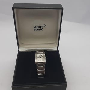 Puede incluir: Un reloj Montblanc plateado con esfera rectangular y correa de acero inoxidable, presentado en una caja negra. La esfera del reloj presenta números romanos y el logotipo de Montblanc. La caja tiene el logotipo de Montblanc.