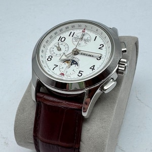 May include: A silver-tone Delma automatic wristwatch with a white dial, black Arabic numerals, and a brown leather strap. The watch features a moon phase indicator, day and month sub-dials, and a date hand pointing to the outer ring of numbers.