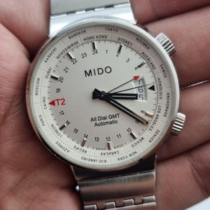 Puede incluir: Reloj Mido All Dial GMT Automatic plateado con esfera blanca. El reloj cuenta con un bisel de hora mundial, una pantalla de fecha y una correa de acero inoxidable. La esfera del reloj muestra la hora en formato de 24 horas.