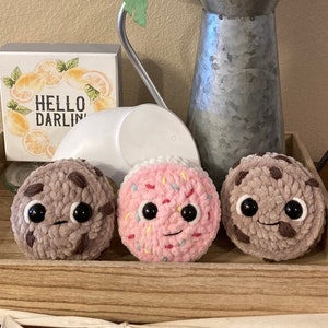 May include: Three crocheted plush cookies on a wooden tray with a white ribbon. The cookies are chocolate chip, pink sprinkle, and chocolate chip. The tray is decorated with a sign that says "Hello Darlin".