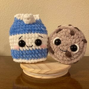 May include: A crocheted milk carton and chocolate chip cookie plush toy with smiling faces and googly eyes. The toys are sitting on a wooden base.