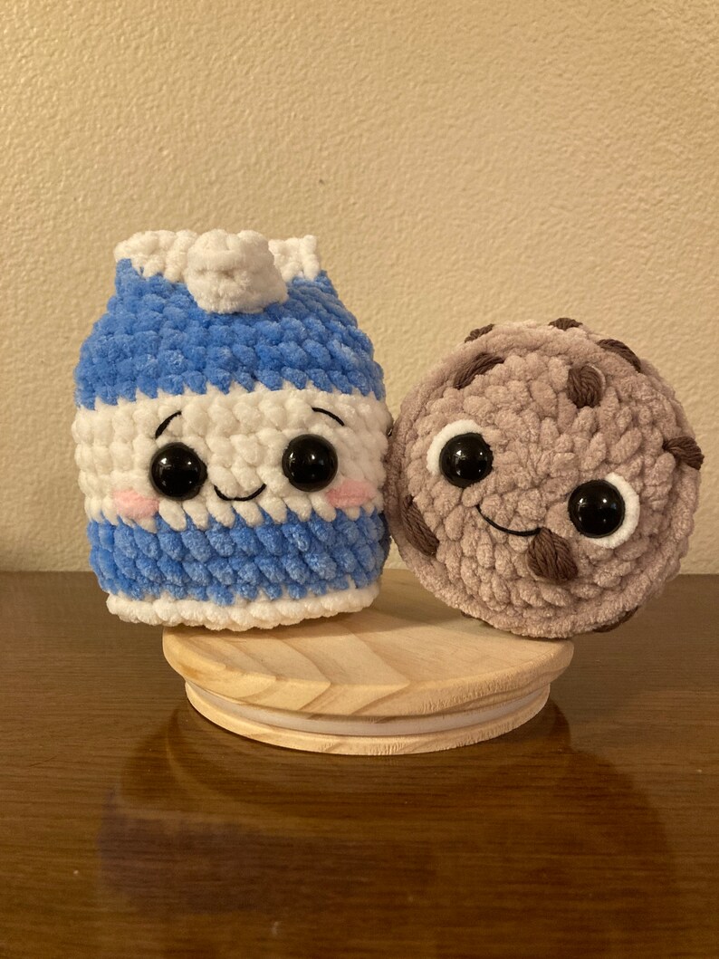 May include: A crocheted milk carton and chocolate chip cookie plush toy with smiling faces and googly eyes. The toys are sitting on a wooden base.