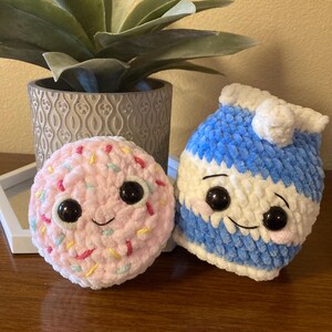 May include: Two crocheted plush toys, one shaped like a donut with pink frosting and colorful sprinkles, the other shaped like a milk carton with a blue and white striped design. Both toys have black eyes and a smiling expression.