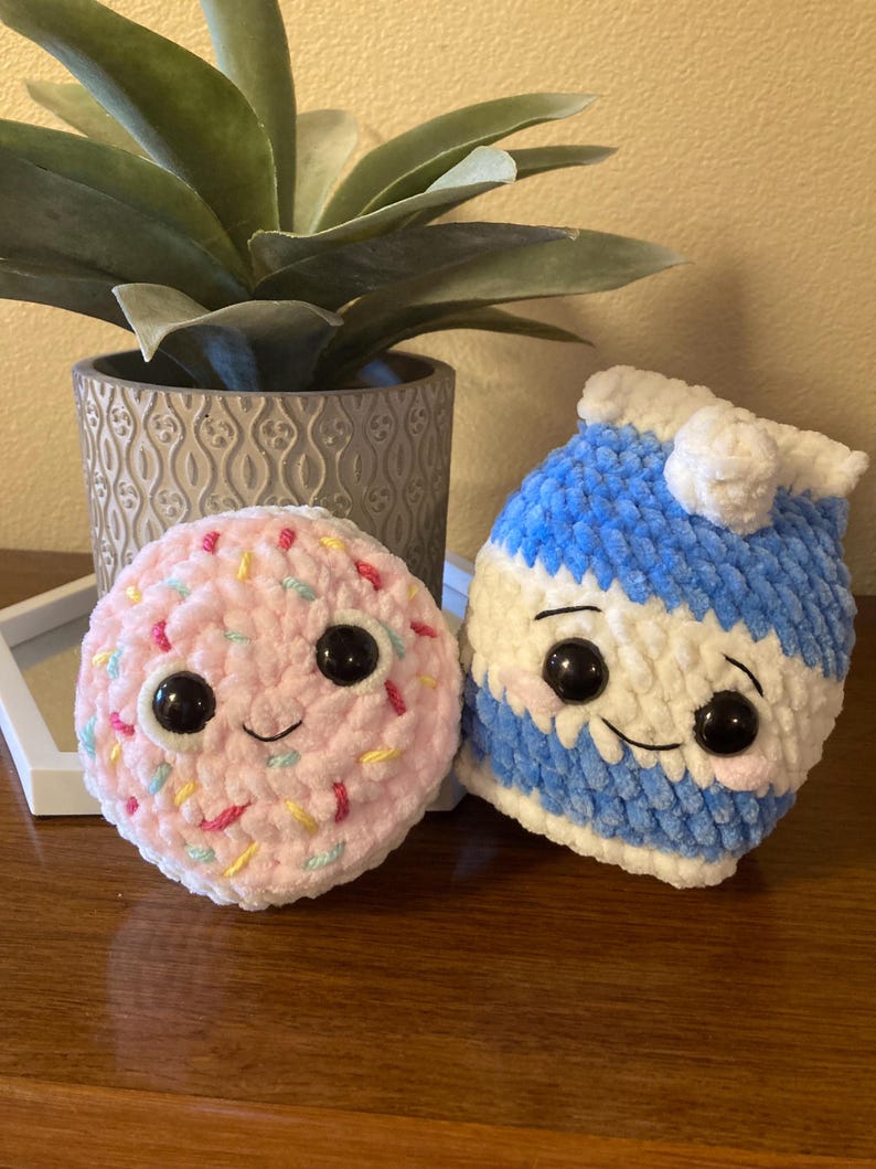 May include: Two crocheted plush toys, one shaped like a donut with pink frosting and colorful sprinkles, the other shaped like a milk carton with a blue and white striped design. Both toys have black eyes and a smiling expression.