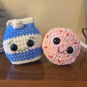 May include: Two crocheted plush toys, one shaped like a milk carton and the other shaped like a donut. The milk carton is blue and white with a white lid and black eyes. The donut is pink with colorful sprinkles and black eyes.