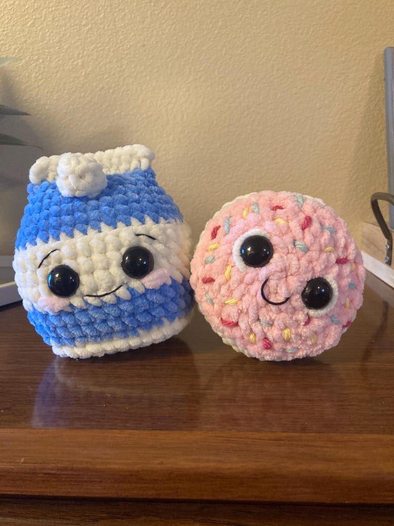 May include: Two crocheted plush toys, one shaped like a milk carton and the other shaped like a donut. The milk carton is blue and white with a white lid and black eyes. The donut is pink with colorful sprinkles and black eyes.