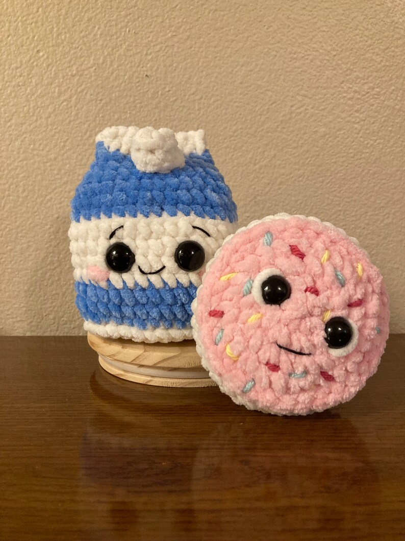 May include: Two crocheted plush toys, a blue and white milk carton and a pink donut with colorful sprinkles. Both toys have black eyes and smiling mouths.