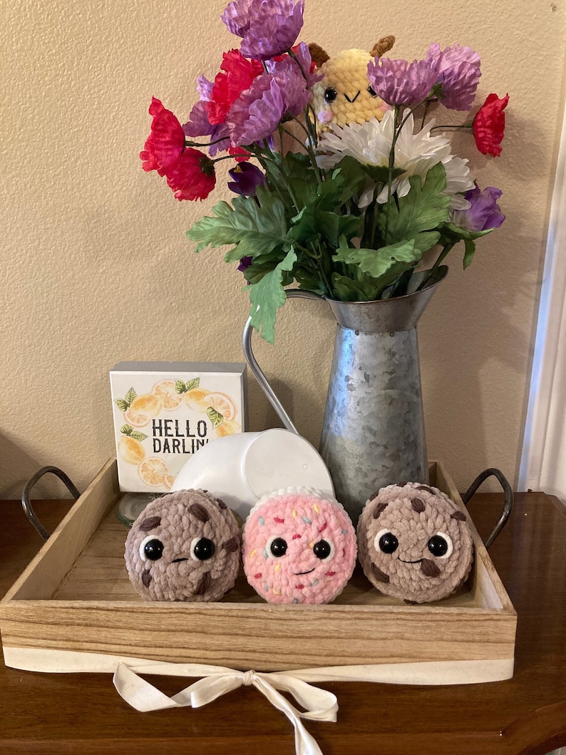 May include: Three crocheted plush cookies on a wooden tray with a white ribbon. The cookies are chocolate chip, pink sprinkle, and chocolate chip. The tray is decorated with a sign that says "Hello Darlin".