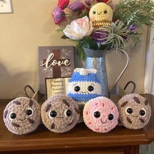 May include: Four crocheted plush toys in the shape of cookies, a blue and white striped milk carton, and a yellow and brown bee. The cookies are brown, pink, and a light brown with chocolate chips. The toys are sitting on a wooden surface with a sign that says "love" in front of them.