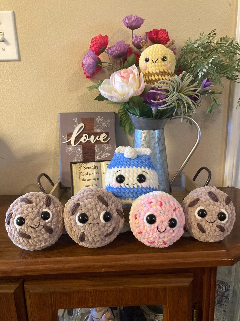 May include: Four crocheted plush toys in the shape of cookies, a blue and white striped milk carton, and a yellow and brown bee. The cookies are brown, pink, and a light brown with chocolate chips. The toys are sitting on a wooden surface with a sign that says "love" in front of them.