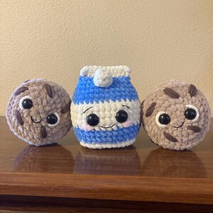 May include: Three crocheted plush toys: a brown chocolate chip cookie, a blue and white milk carton, and another brown chocolate chip cookie. Each toy has a smiling face with black eyes.
