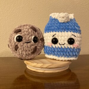 May include: A crocheted milk carton and chocolate chip cookie plush toy with smiling faces and big eyes. The toys are sitting on a wooden base.