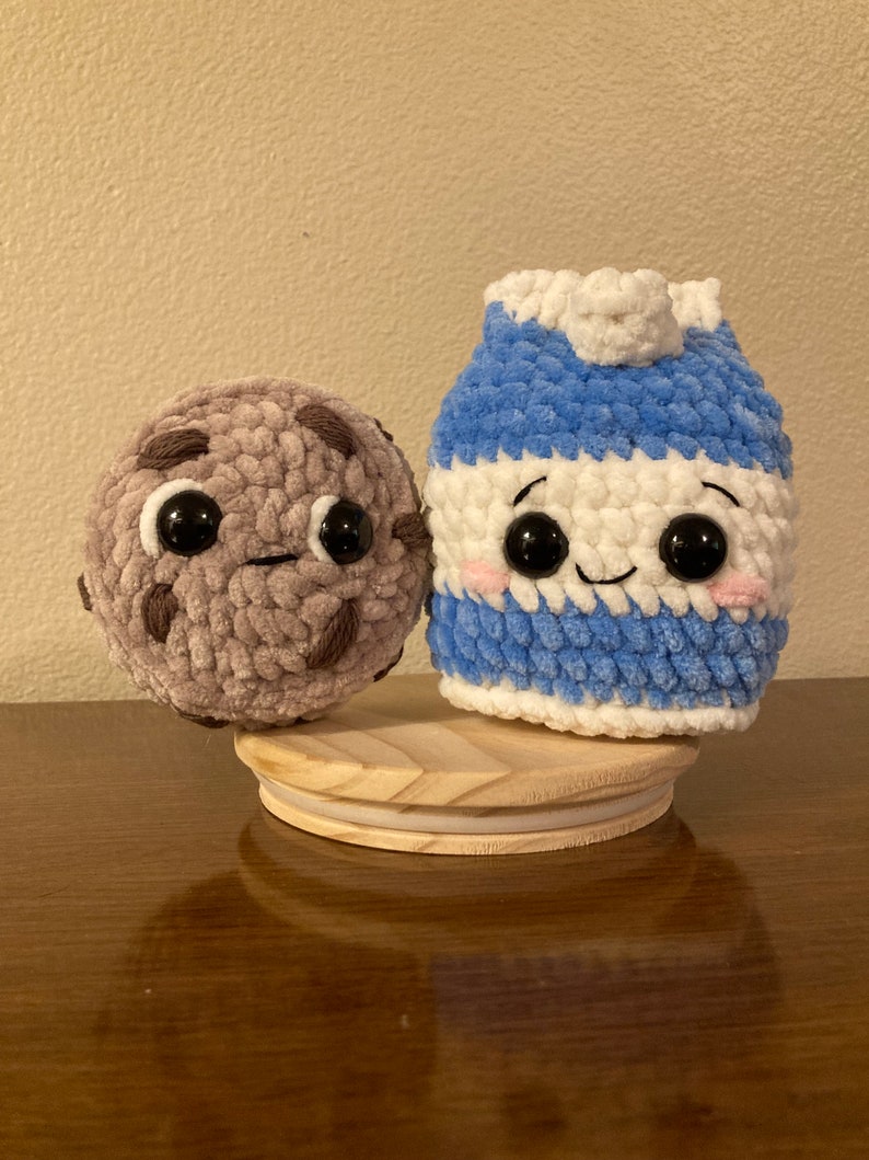 May include: A crocheted milk carton and chocolate chip cookie plush toy with smiling faces and big eyes. The toys are sitting on a wooden base.