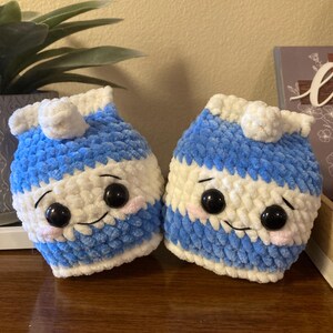 May include: Two crocheted milk carton plushies with smiling faces. The plushies are blue and white with black eyes and pink cheeks.