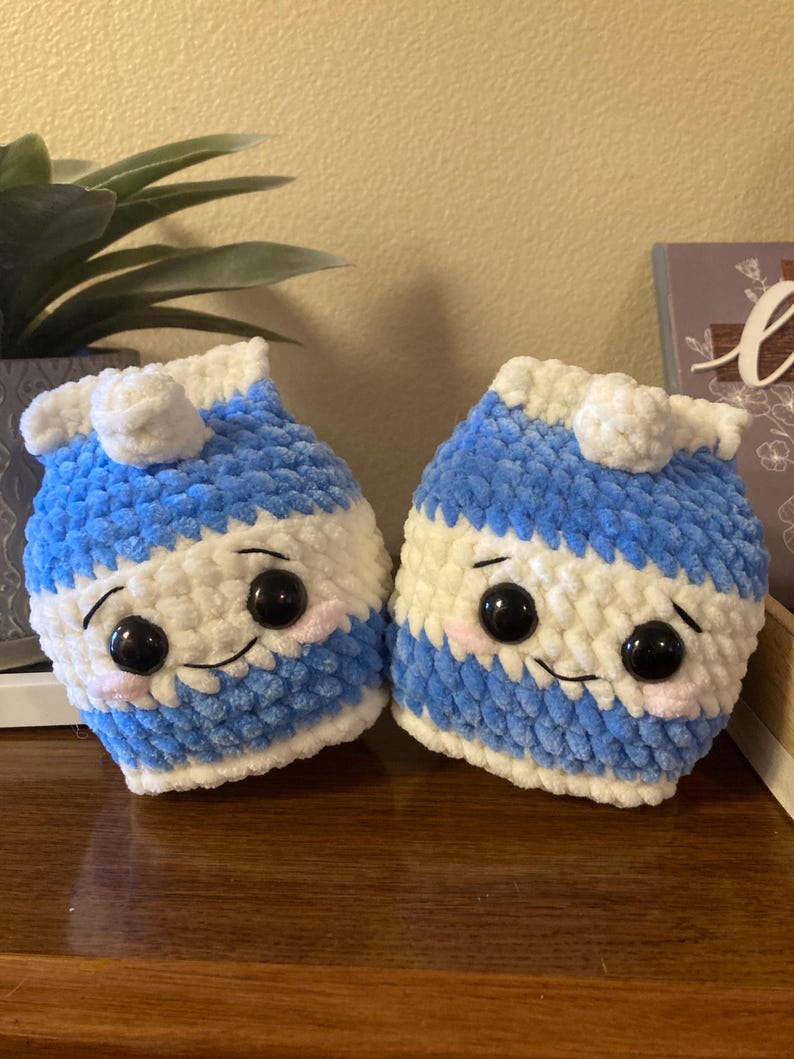 May include: Two crocheted milk carton plushies with smiling faces. The plushies are blue and white with black eyes and pink cheeks.