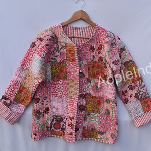 May include: A patchwork jacket in shades of pink, featuring floral and geometric patterns. The jacket has a V-neck, long sleeves, and a quilted texture. The sleeves and edges are trimmed with a striped fabric.