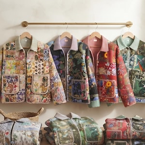 May include: Four quilted jackets with patchwork patterns in various colors hang on a gold rod. Each jacket features a collared neckline, long sleeves, and two front pockets. Three additional folded jackets are stacked on a wooden surface below.