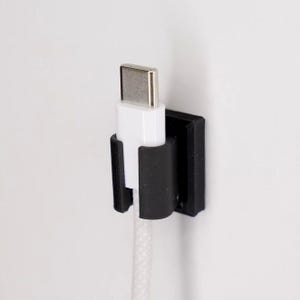 May include: A black wall-mounted charging cable holder with a white charging cable and silver connector. The holder is designed to keep the cable tidy and accessible. The cable is white with a braided texture.