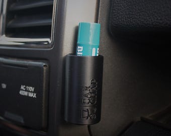 Op de afbeelding: Een matzwarte cilindrische houder gemonteerd op het dashboard van een voertuig, met de in reliëf aangebrachte tekst CAR CHAP. Een blauwgroene lippenbalsemstift zit in de houder, die naast een ventilatierooster en een stopcontact is geplaatst.