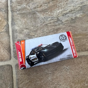 May include: A black and white toy police car with a red and blue light bar, displayed on its original packaging. The packaging reads "MBX Rescue" and "65 Anniversary". The toy car is a '14 Corvette Stingray.