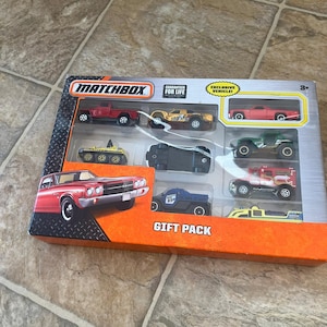 May include: A Matchbox gift pack containing several toy cars in a silver and orange box. The set includes a red pickup truck, a yellow race car, and a red sports car. The box is labelled "GIFT PACK" and "GUARANTEED FOR LIFE".