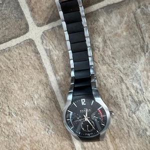 May include: A Skagen watch with a black face and silver-tone stainless steel strap. The watch has a black dial with silver hands and markers, and a date display. The watch is on a tiled surface.