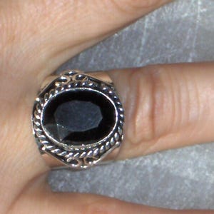 May include: A silver ring with an oval, black gemstone. The ring has a decorative, rope-like border around the gemstone and a detailed design on the band. The ring is shown on a finger.