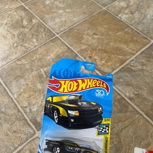 May include: A black and yellow Hot Wheels toy car on a blue and white card. The car has the text "MOONEYES" and "GOOD YEAR" printed on it. The card reads "Hot Wheels 50".