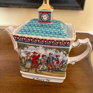 Könnte beinhalten: Eine dekorative Teekanne mit Golfmotiv. Der Deckel ist ein türkisblaues Dach mit einer goldenen Uhrturmspitze. Der Körper zeigt eine gemalte Szene von Golfspielern und den Text "A Round of Golf".