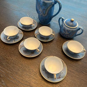 Op de afbeelding: Een blauwe theeset met een drakenontwerp. De set bevat een hoge theepot, een kleine theepot en zes kopjes met schotels. De kopjes en schotels hebben een gouden rand. De set staat op een houten tafel.
