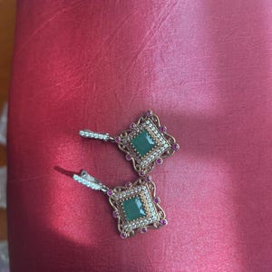 May include: A pair of ornate earrings with a square emerald-green center stone. The earrings have a gold-colored filigree frame, accented with small pink gemstones and clear crystals. The earrings are set against a red background.