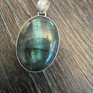 May include: Oval labradorite pendant with a silver-colored bail. The gemstone has a dark teal and green iridescence. The pendant is set in a silver-colored bezel. The pendant is displayed on a brown wooden surface.