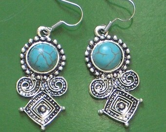May include: A pair of silver-toned earrings with turquoise-colored stone centers. The earrings have a decorative design with a round top, a swirl design, and a diamond-shaped bottom. The background is a solid green color.