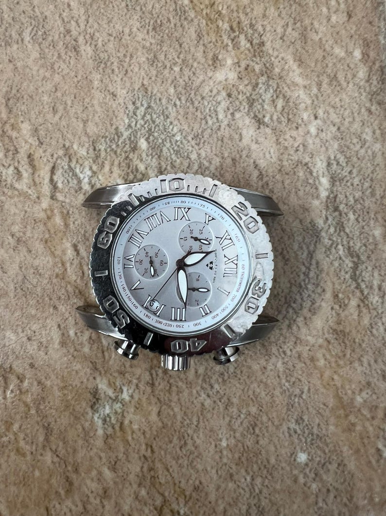May include: Silver-toned wristwatch with a white face and three sub-dials. The watch features Roman numeral hour markers and a textured bezel with minute markings. The watch has a silver band and a crown for adjusting the time.