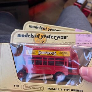 May include: A red Matchbox 1922 A.E.C. 'S' Type Omnibus model, featuring the Schweppes logo. The model is in its original packaging, labeled "models of yesteryear". The packaging includes a price tag.