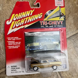 Peut inclure: Modèle réduit Johnny Lightning d'une Chevy Nomad de 1956. La voiture est dorée et beige, avec un moteur et un intérieur détaillés. L'emballage est rouge et porte l'inscription "TRI-CHEVY" et "1956 CHEVY NOMAD".