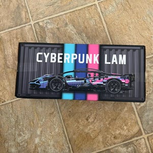 Könnte beinhalten: Eine schwarze Box mit einem futuristischen Sportwagendesign in Blau und Pink. Der Text "CYBERPUNK LAM" ist weiß. Das Auto hat schwarze Räder und ein niedriges Profil. Es handelt sich wahrscheinlich um einen Modellbausatz.