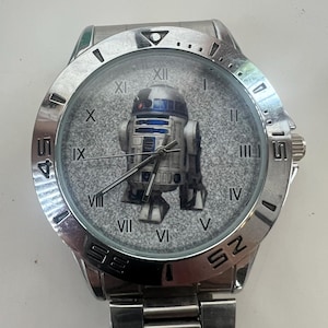 May include: A silver-toned wristwatch featuring the iconic R2-D2 droid from Star Wars on the watch face. The watch has a stainless steel band and a round face with Roman numeral hour markers. The watch hands are black.