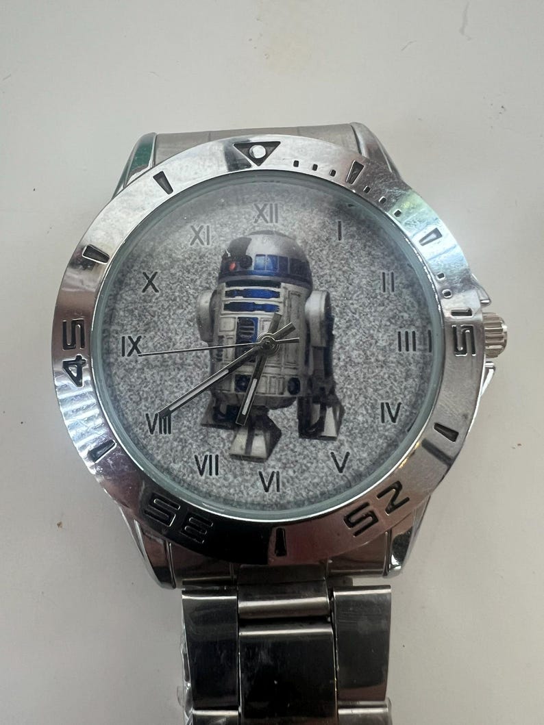 May include: A silver-toned wristwatch featuring the iconic R2-D2 droid from Star Wars on the watch face. The watch has a stainless steel band and a round face with Roman numeral hour markers. The watch hands are black.