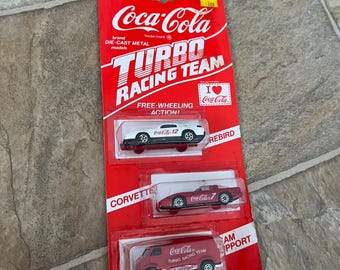 May include: A red Coca-Cola Turbo Racing Team die-cast metal model set. The set includes three miniature vehicles: a white car, a red Corvette, and a red van. The packaging features the Coca-Cola logo and the words "Turbo Racing Team".