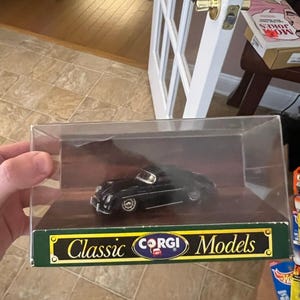 May include: A black Corgi Classic Models car in a clear display case. The model car is a vintage design with silver wheels. The display box has a green and gold label with the words "Classic Models" and the Corgi logo.