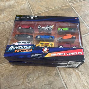 May include: A nine-pack of Adventure Force die-cast vehicles in a clear plastic package. The set includes various car models in red, green, orange, blue, and black. The package has the Adventure Force logo and the text "DIE-CAST VEHICLES".