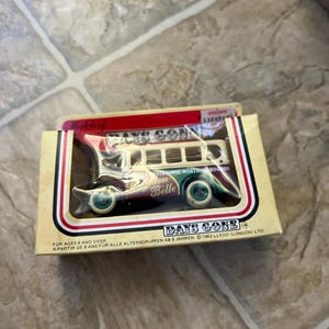 Könnte beinhalten: Ein Days Gone Modellbus in der Originalverpackung. Der Bus ist weiß mit roten und grünen Akzenten, und die Box enthält Text auf Englisch und Deutsch. Das Modell ist ein "Brighton Belle" Bus.
